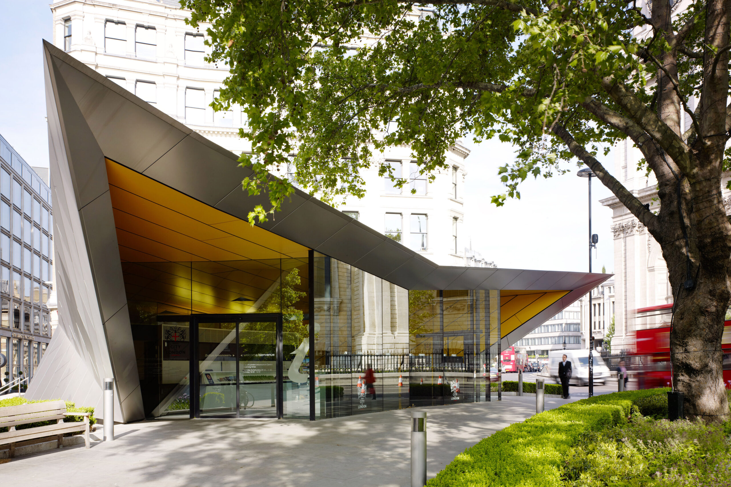Exterior view of the City Information Centre by St. Paul's Cathedral, featuring a modern angular design with glass walls under a large, sloping metal roof. A red bus passes by on the adjacent street, and bare trees and a wooden bench are visible in the foreground.