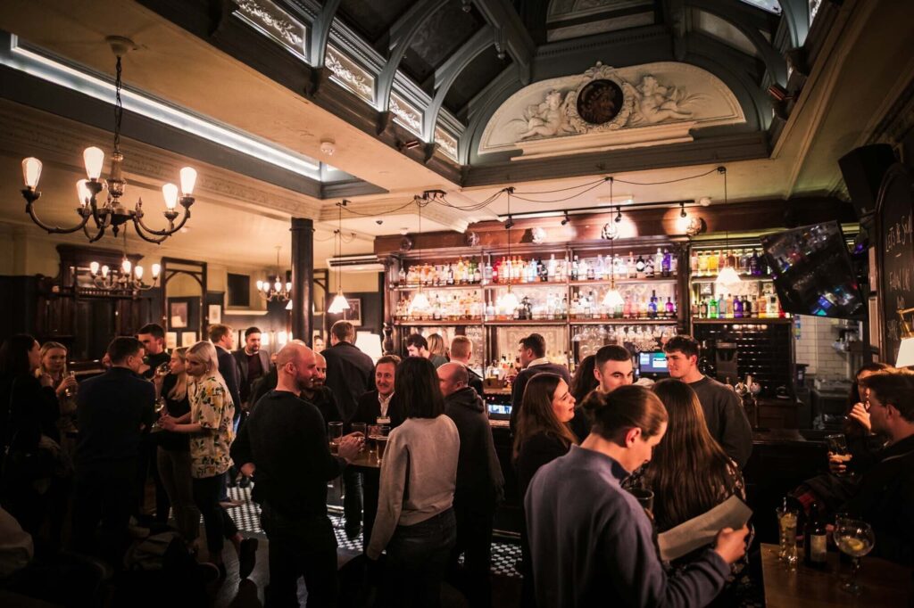 9 Best Pubs in the City of London -  The punch Tavern pub interior, with a crowd of people gathered round a old school bar drinking and having a nice time in the evening.