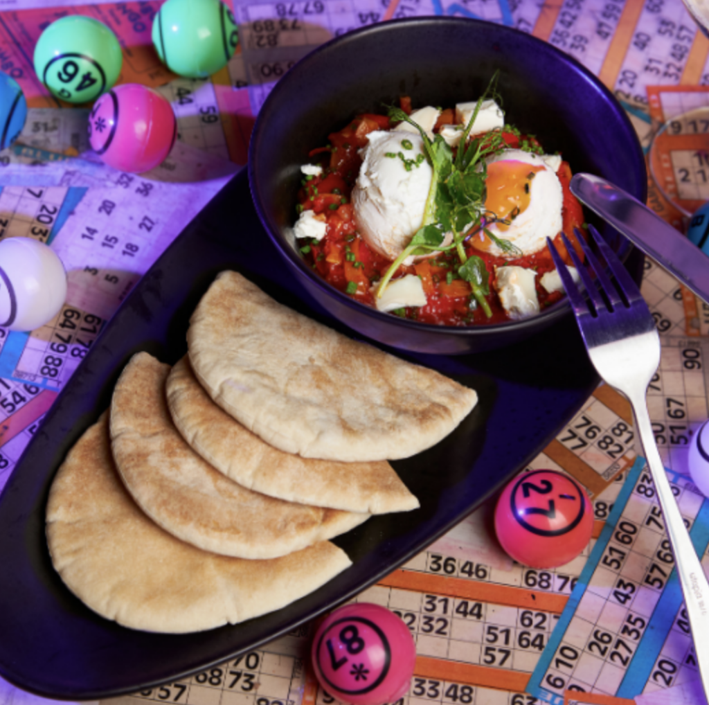9 Best Brunch Spots in the City of London - Dabbers Bingo - table covered in bingo cards and balls with a plate of pitta and a bowl of Shakshouka and poached eggs 