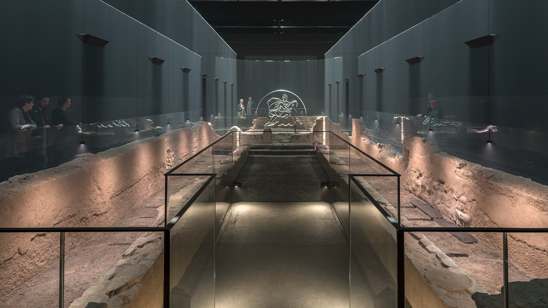 Evening in the City - Temple Mithras – interior of exhibition space, low stone foundation ruins lit with down lamps in a long room of grey walls