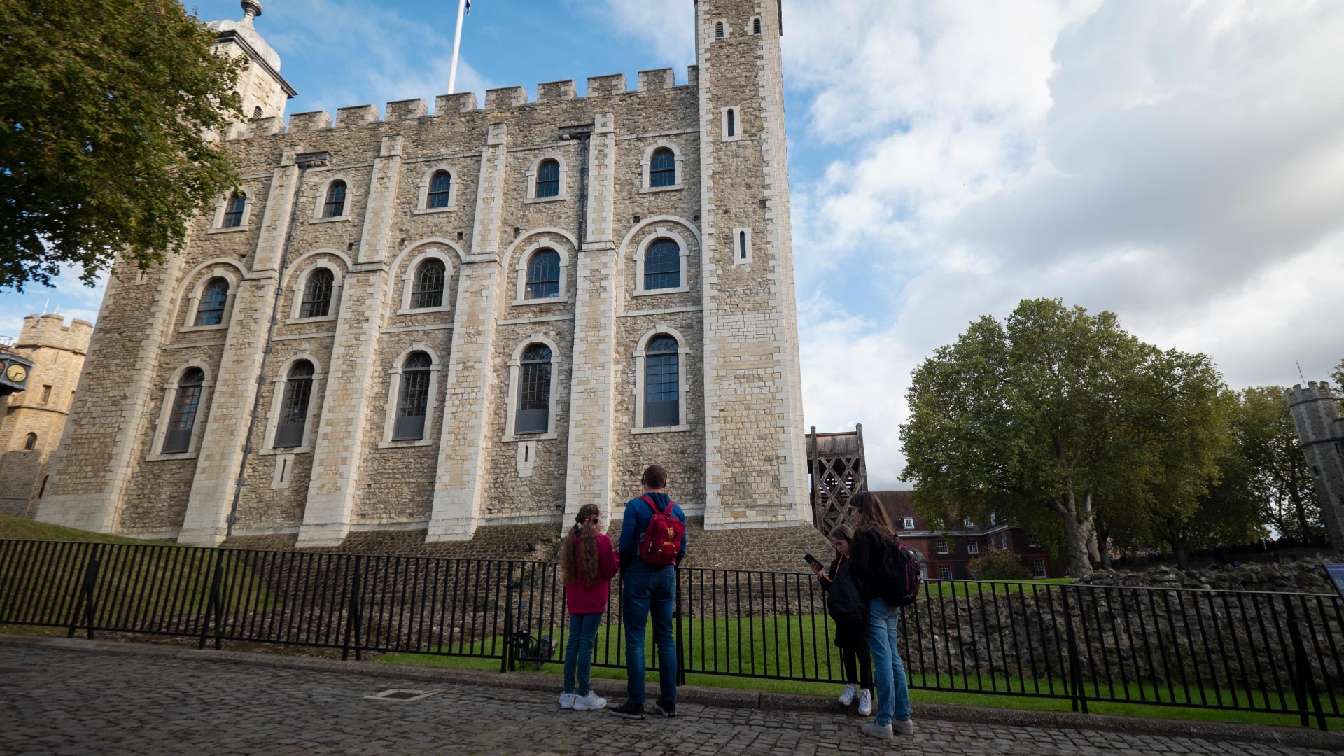 Family Fun Activities to Enjoy on the Weekend in the City of London - A picture of the Tower of London on a summer day, behind the fence there is a family of four standing and listening to an audio guide, an older male, and thee younger girls.
