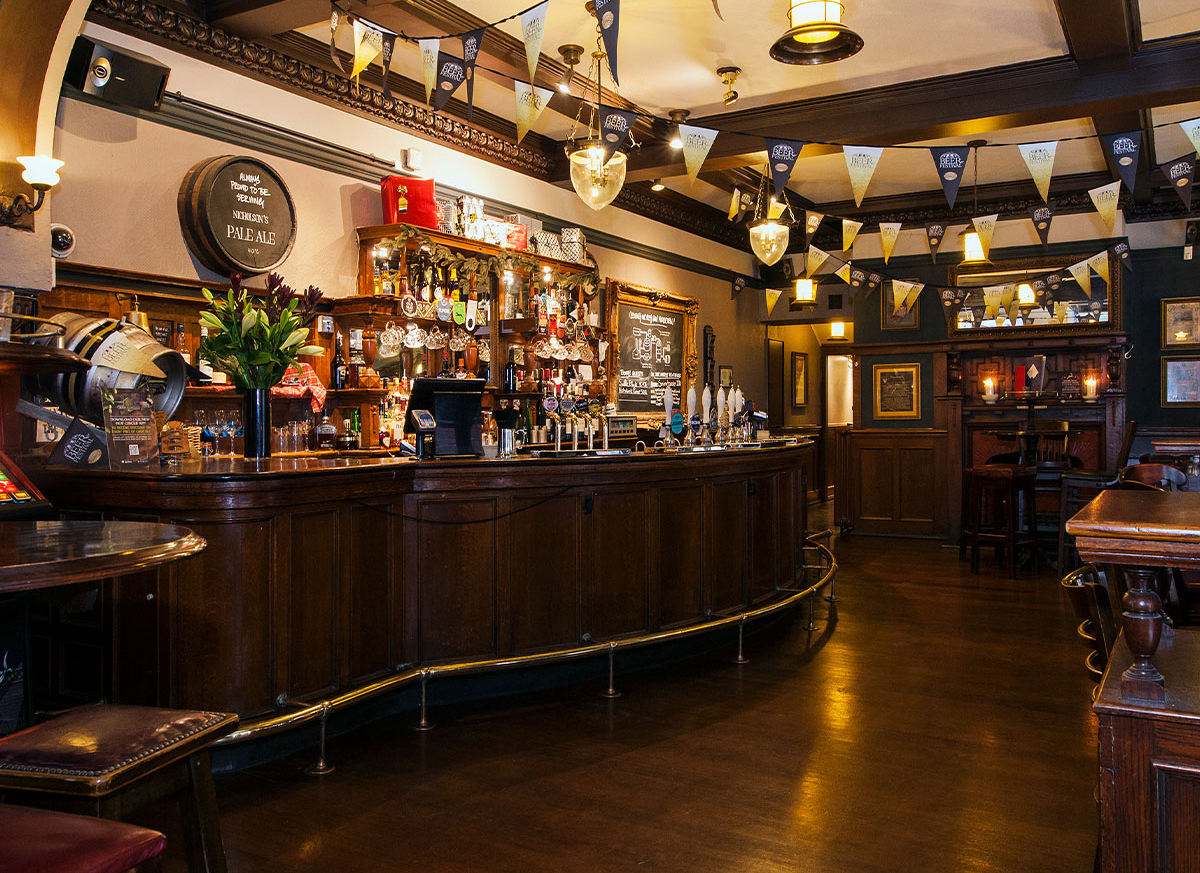 9 Best Pubs in the City of London - Williamson’s Tavern interior with a curved bar in an old english style. There are flags going across the ceiling.