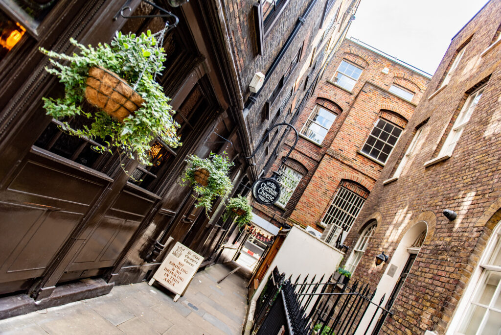 9 Best Pubs in the City of London - A slanted view of the Ye Olde Cheshire Cheese pub, looking down an narrow alley leading on to the road, with a hanging plant basket in the foreground 