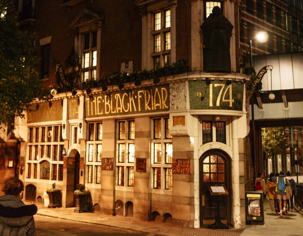 9 Best Pubs in the City of London - The Black Friar pub at night, a narraow building with 'The Black Friar' written in mosaics on the side, with a human-sized friar standing over the door frame entrance.