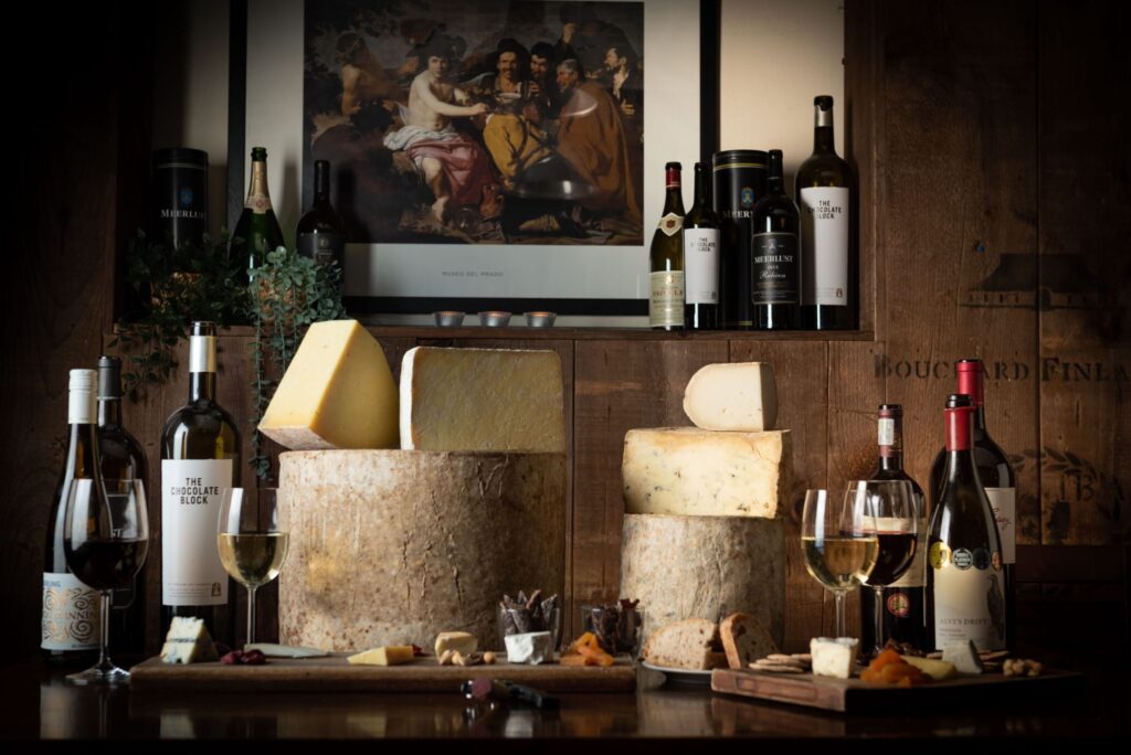 19 best restaurants to eat in the City of London - Vivat Bacchus - table of bottle and glasses of white and red wine, cheese boards and large full and quarter sections of cheese wheels in the background