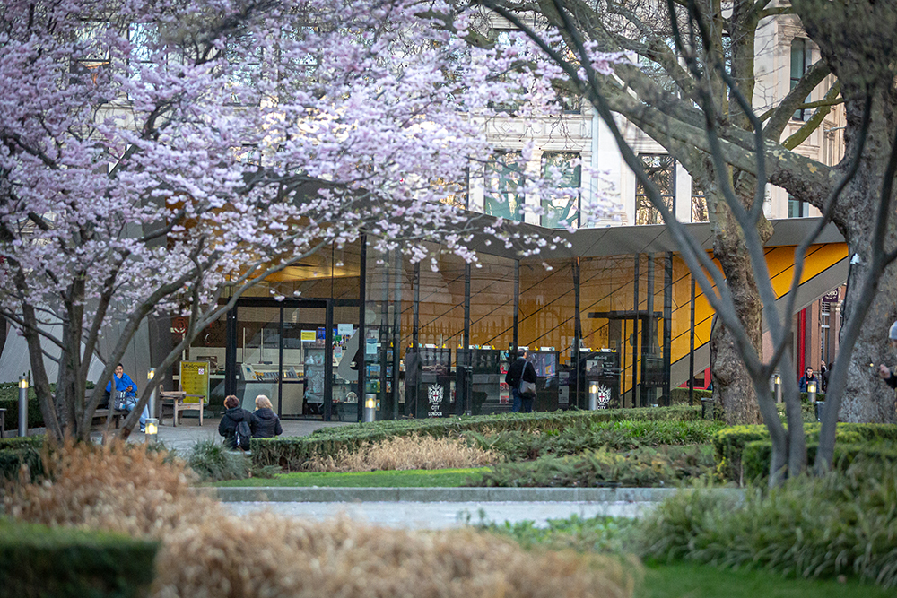 Family Fun Activities to Enjoy on the Weekend in the City of London - The City Centre is a modern building with a yellow interior, and we are viewing it through light pink blossom on near by trees with the green gardens in the foreground.