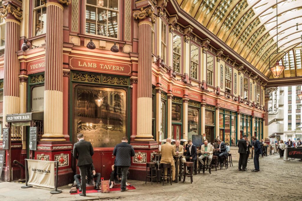 9 Best Pubs in the City of London - The Lamb Tavern in Leadenhall market. Leadenhall market is a red outdoor covered market, with ornate pillars. Outside the Lamb tavern there are tables where people are drinking as well as traditional shoe shiners.
