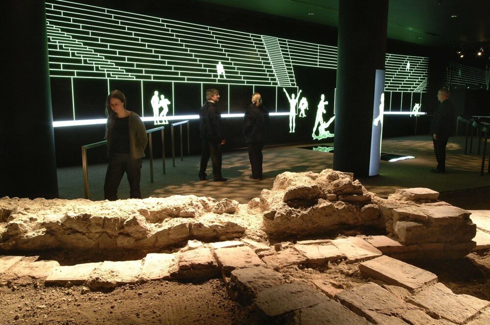 Visitors looking at Roman ruins in London's Roman Amphitheatre with lights simulating gladiator fights in the background