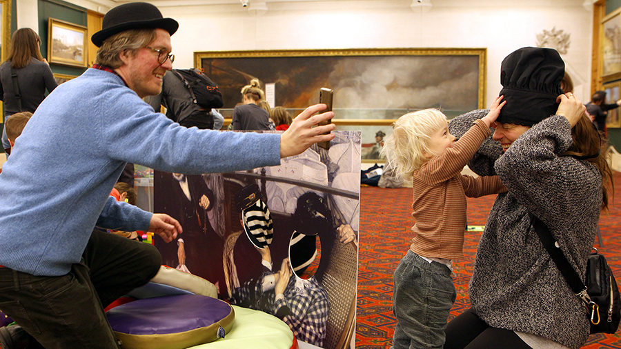Toddler trying to take the hat from his mum's head at an art gallery and man taking a photo of them