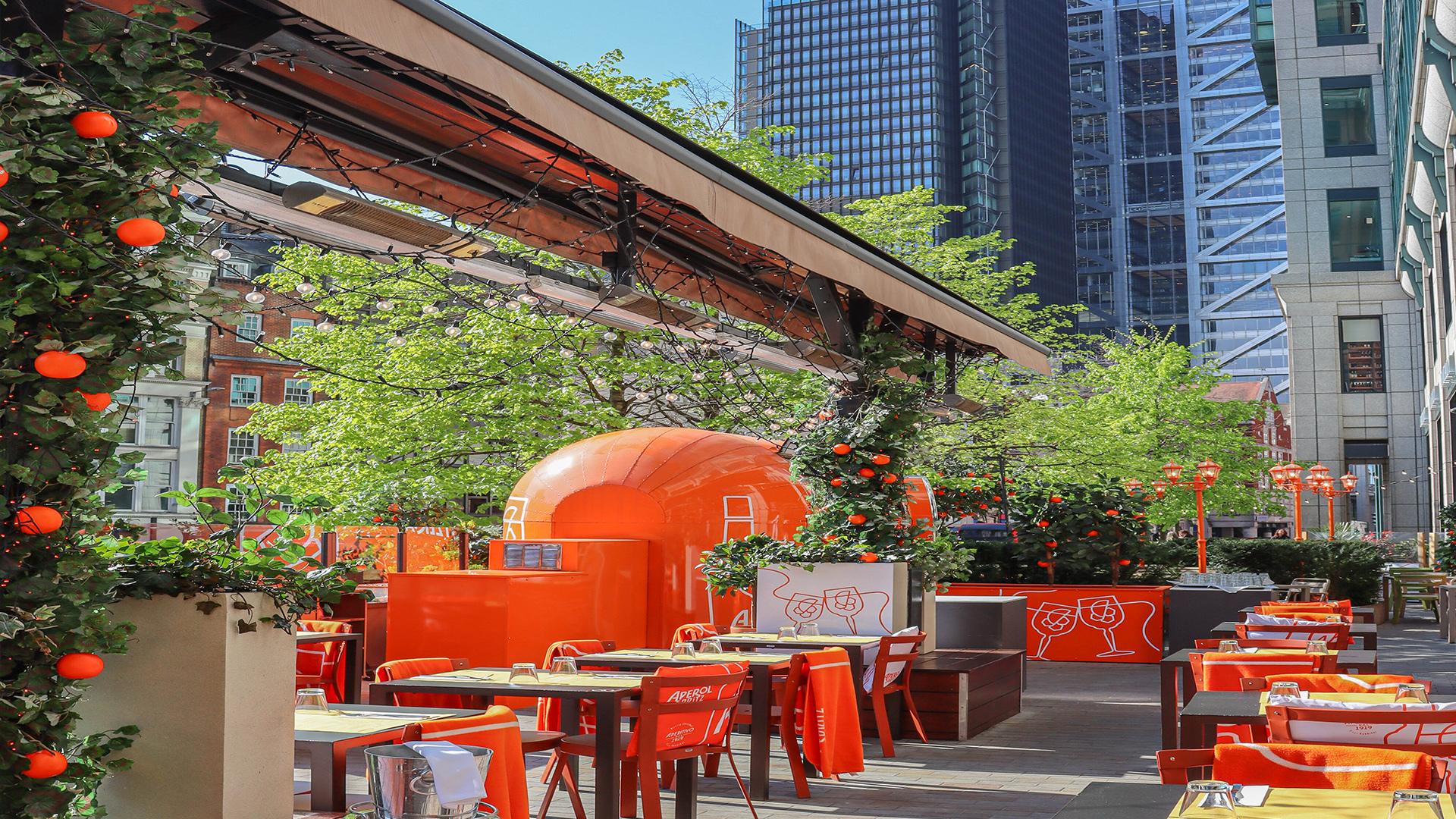 Eataly's outdoor terrace in the City of London - with orange chairs, orange lights and ornamental orange trees. Green trees and modern buildings in the background.