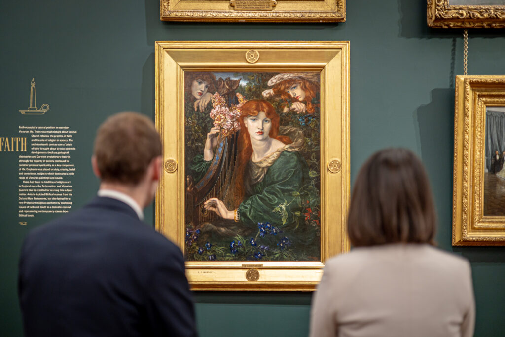 Two people looking at a golden framed painting of a woman with red hair wearing a green gown, hanging on a dark green wall
