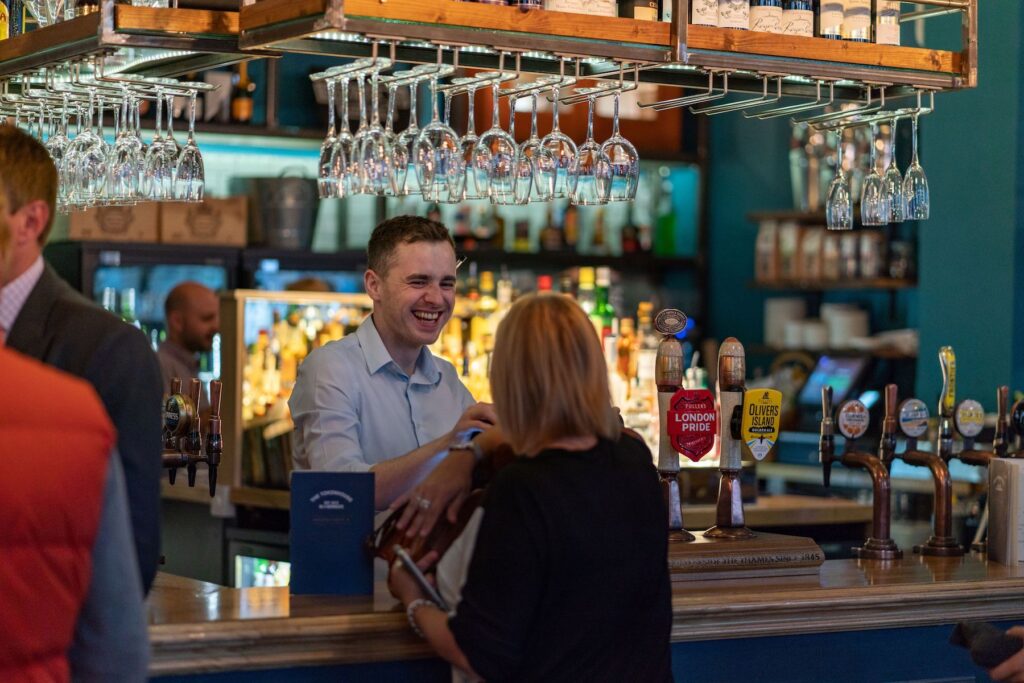 The Tokenhouse - barman smiling while serving a woman at the bar - wine glasses hand from the shelf above