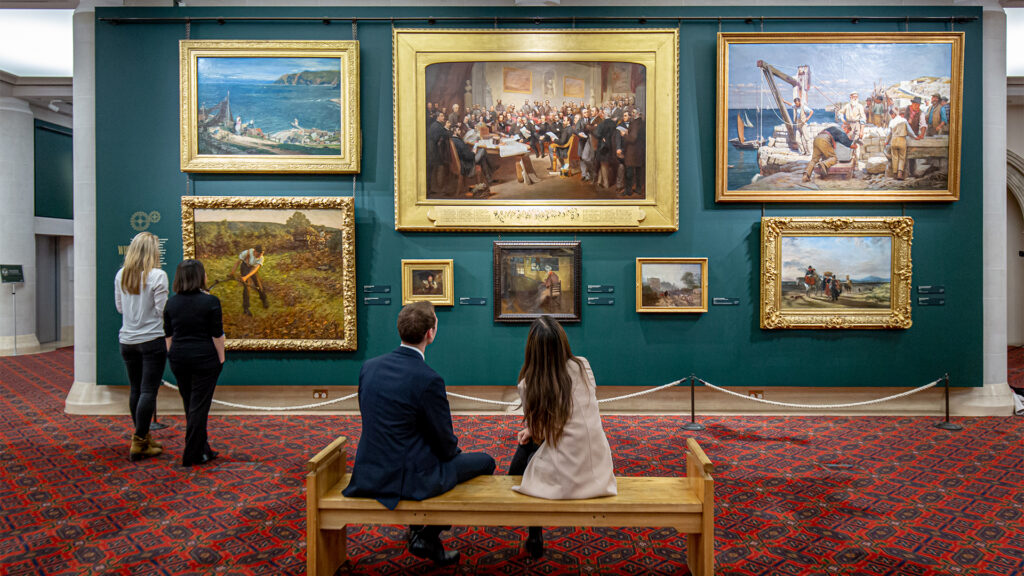 People looking at a selection of paintings with golden frames hanging on a dark green wall