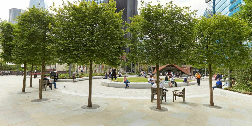 Aldgate Square in the City of London. Green trees, a lawn and people sitting and enjoying the sunshine.