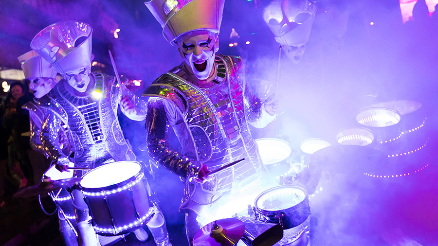 Performers dressed in silver outfits playing drums in a stage illuminated with colourful led lights