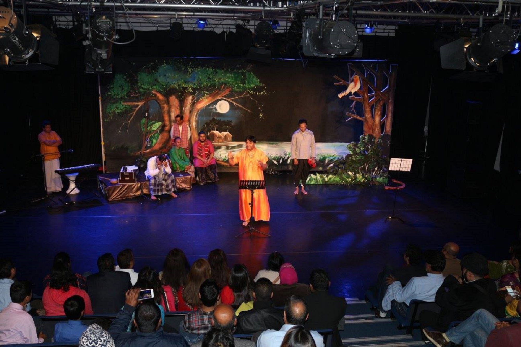 man dressed in yellow clothes on a stage decorated with trees talking to an audience at a theatre
