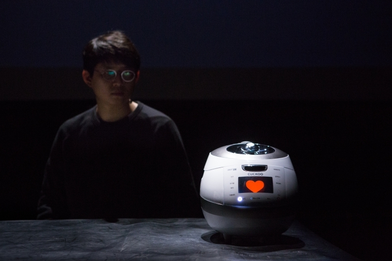 Man looking at a robot with an orange heart on a small digital screen, placed on a table on a black background