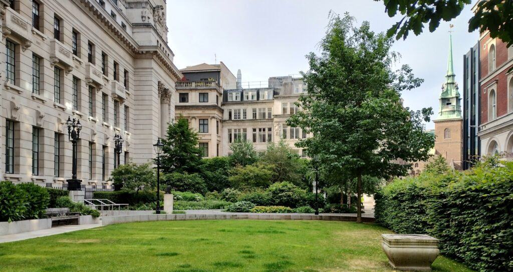 Seething Lane Garden - a large lawn area surrounded by green plants and trees. Left hand side is 10 Trinity Square hotel, in the distance is the steeple of All Hallows by the Tower.
