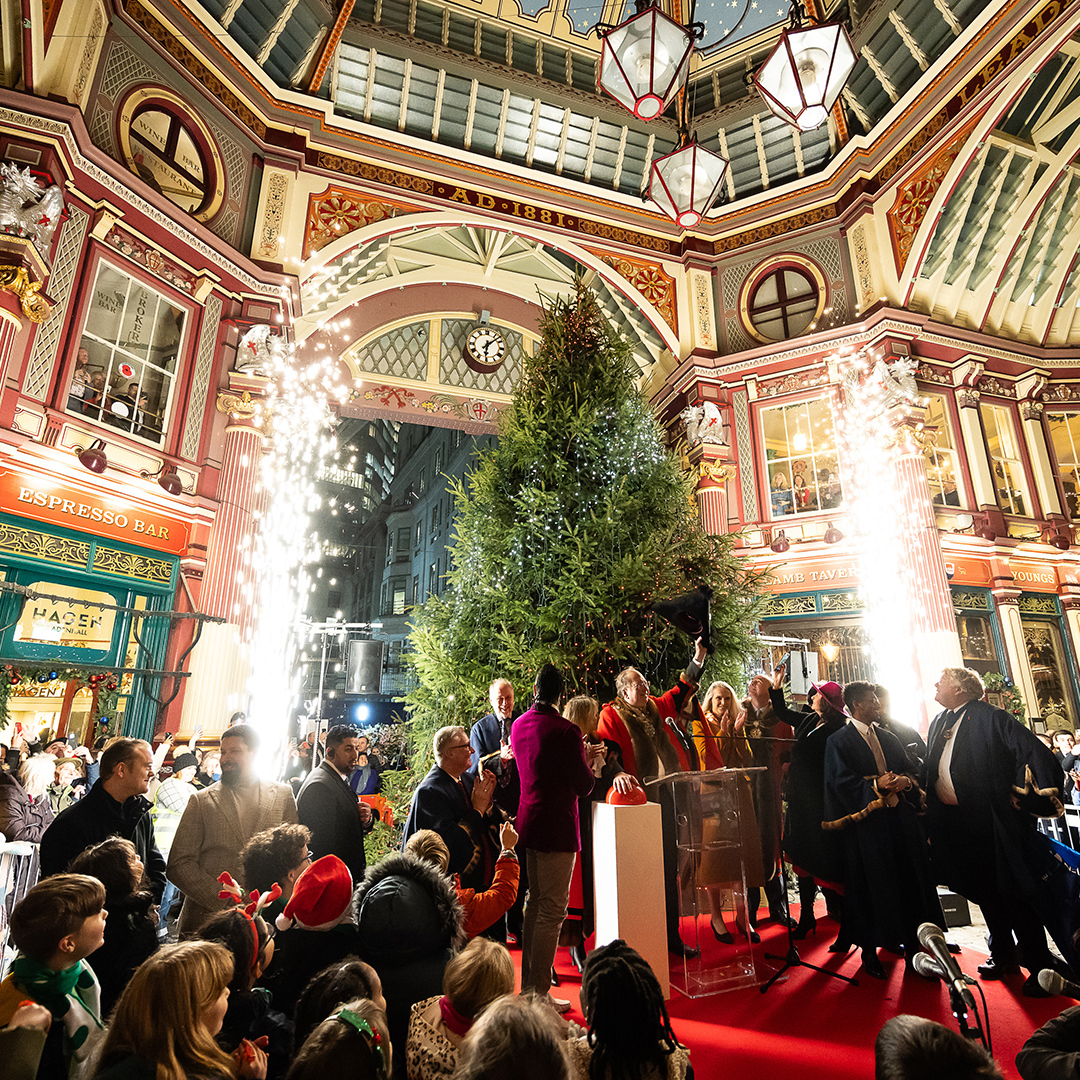 Crowd looking at a group of people standing in front of a Christmas tree in a Victorian market. There are firework fountains at both sides of the tree.