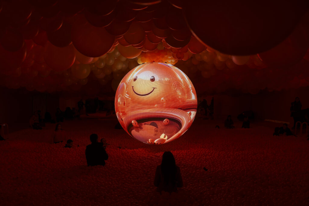 Illuminated red balloon with a projection of a smiley face. The illuminated balloon is hanging from the ceiling in a room covered in thousands of balloons with people in it as well.