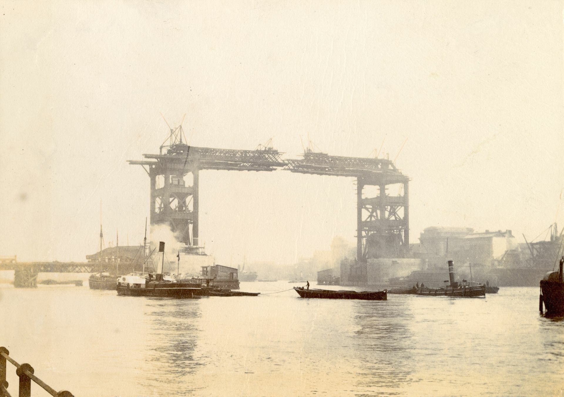 A historic photo of Tower Bridge under construction, showing steamboats on the Thames river and scaffolding around towers