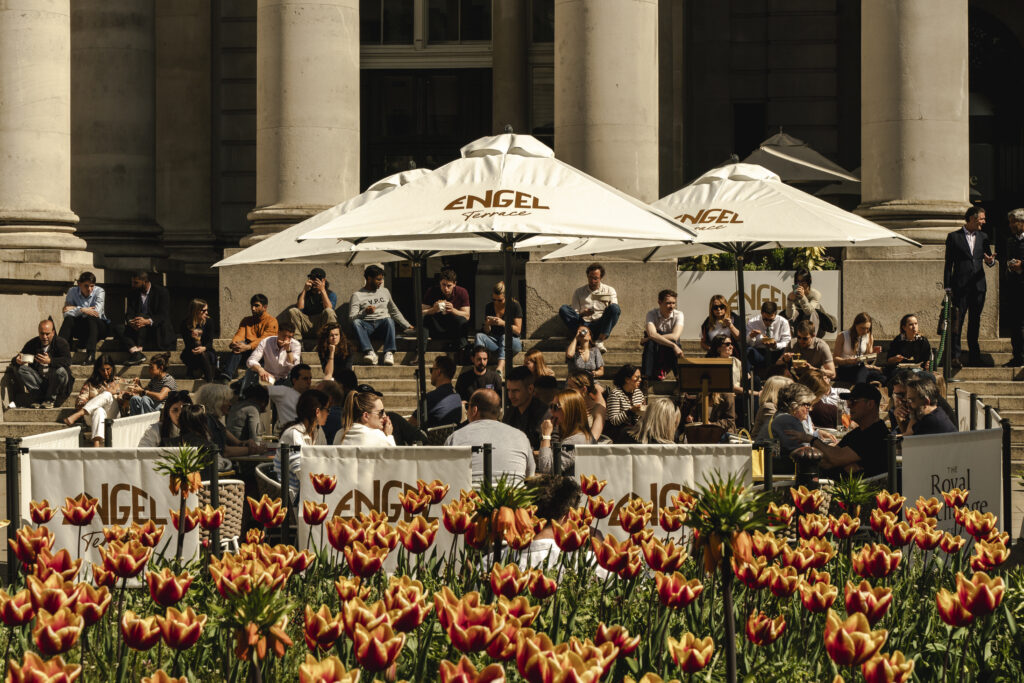 Outdoor seating area with people seated under umbrellas branded with "ENGEL," located near a building with columns, surrounded by vibrant tulips in the foreground.