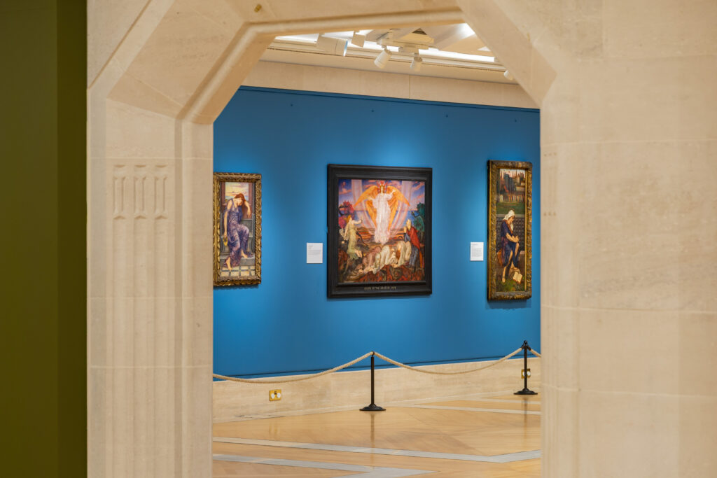Interior view of an art gallery with three framed paintings displayed on a vibrant blue wall, visible through an archway.