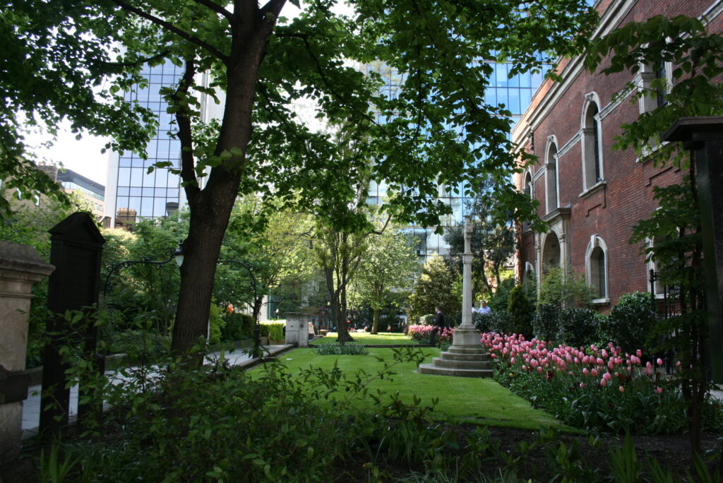 St Botolph’s large churchyard  with green lawn, tall trees, statue of a cross and pink peonies.