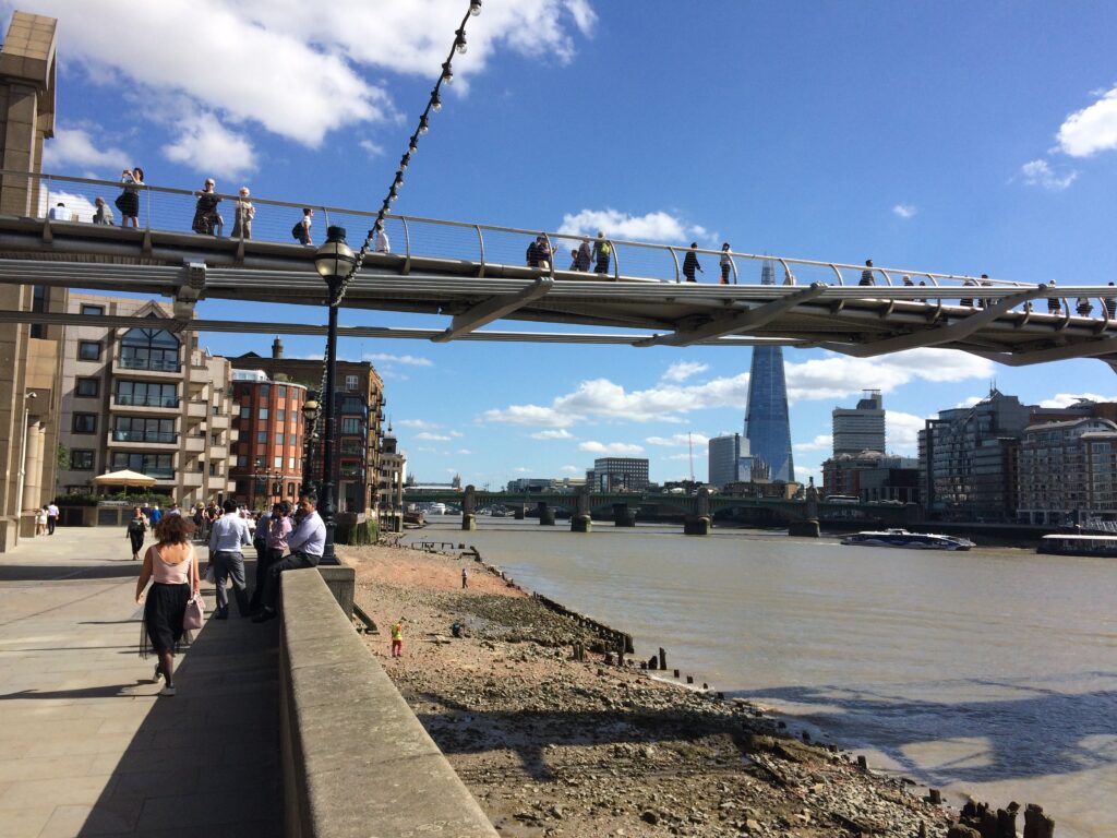 View of Thames path with River Thames on the right, Millenium Bridge can be seen with people walking across it. Blue skies.