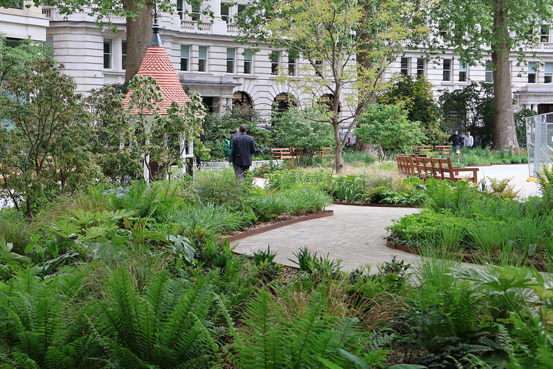 View of a landscaped garden with lush greenery and pathways, featuring benches and a background of classic architectural buildings.