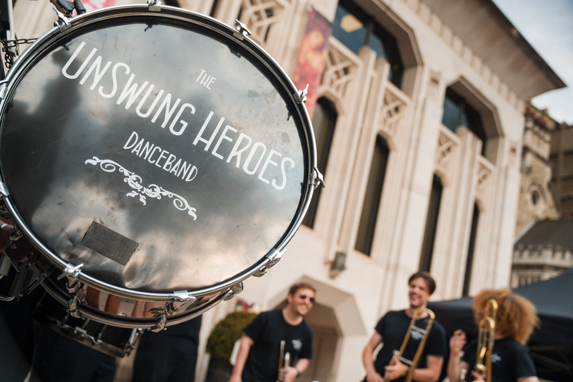 Close up on a dram with wording 'The Unswung Heroes Danceband' with musicians and a historic building in the background