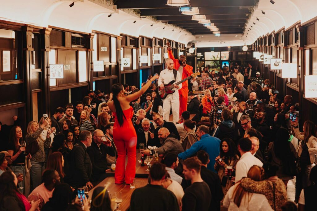 Busy restaurant scene with patrons enjoying live music performed by a guitarist in a vibrant outfit, surrounded by diners engaged in conversation and dining.