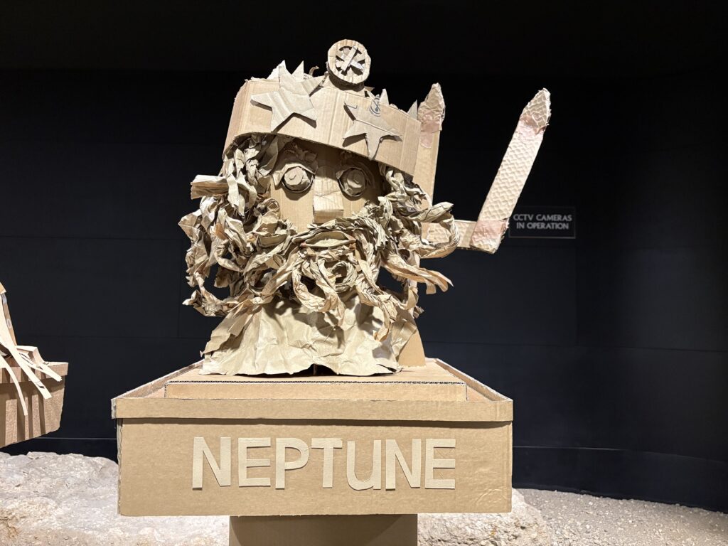 An intricate cardboard sculpture labelled "NEPTUNE," featuring a detailed face with expressive eyes and a beard, adorned with a trident and a sea-themed crown.