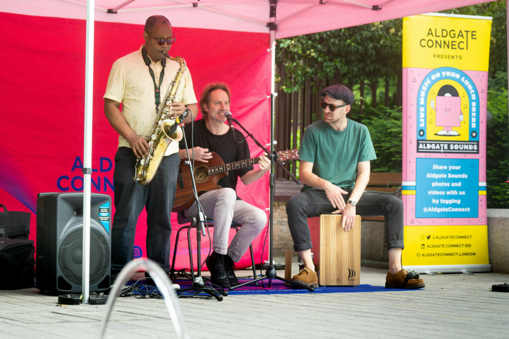 Three individuals are on stage: one is playing a saxophone, another a guitar, and the third a box. They are situated under a red tent.