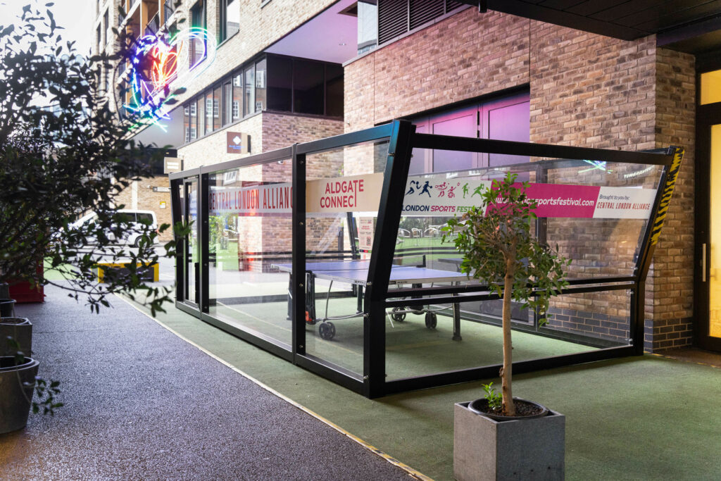 Exterior view of a Padel Table Tennis court at Tower Suits, a modern facility with a glass enclosure and neon signage, featuring table tennis tables inside.