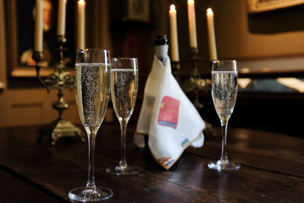 Two champagne flutes filled with sparkling wine, with a bottle of champagne wrapped in a white napkin, set on a wooden table in a dimly lit room with candlelight in the background.