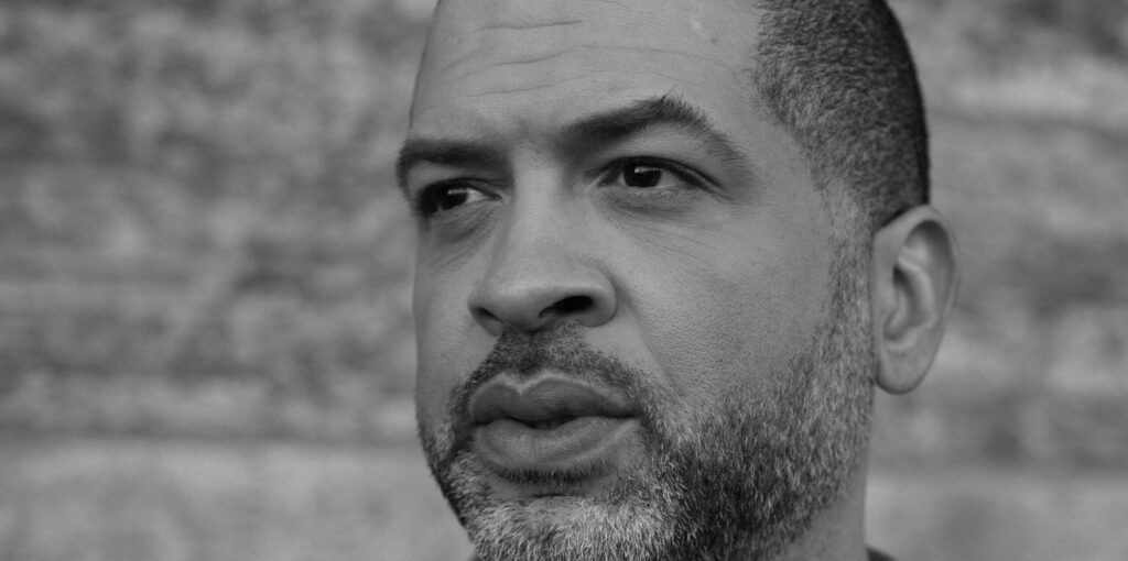 Close-up black and white portrait of an individual with a light beard, looking away from the camera with a thoughtful expression, set against a brick wall background.