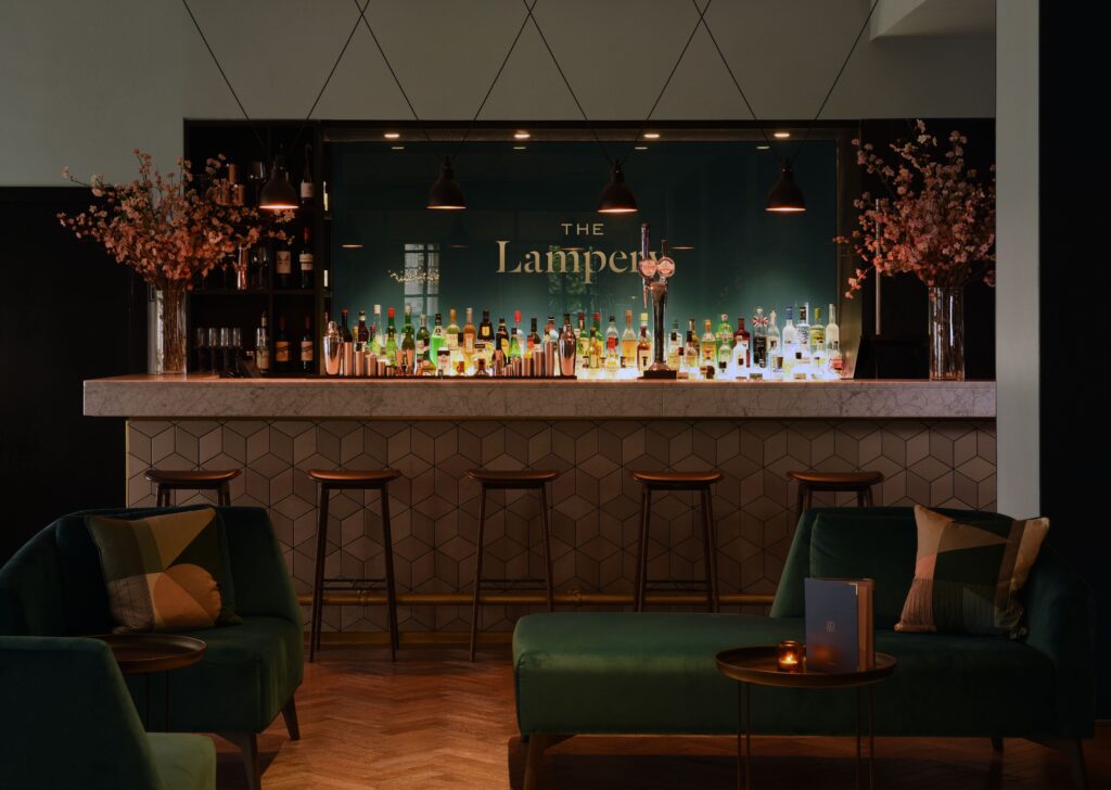 Elegant bar setting at The Lampery, featuring a well-stocked bar counter lined with a variety of spirits, dark stools, ambient lighting, and plush green armchairs surrounded by pink flowering branches.