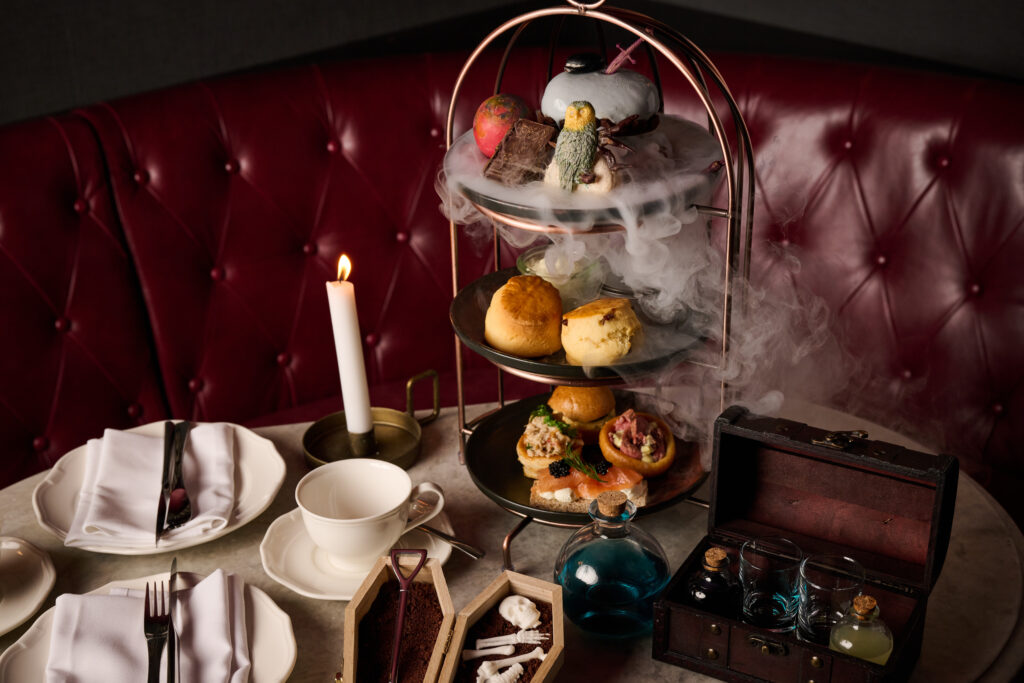 An elegant dining setup featuring a three-tiered serving tray filled with assorted desserts and sandwiches, surrounded by a mist of dry ice. The tray is on a table set with fine china and silverware, illuminated by candlelight, against a backdrop of a red tufted booth.