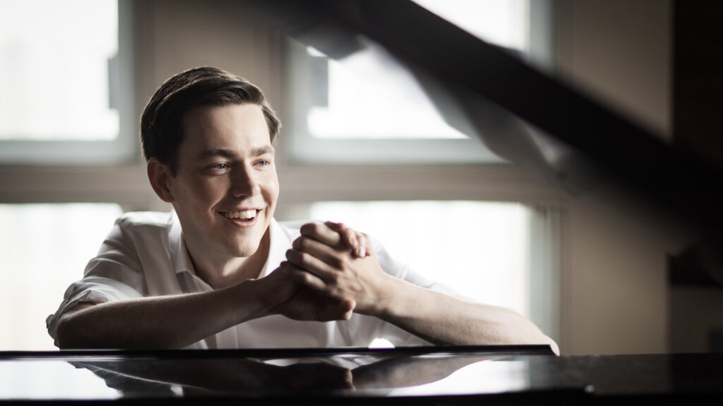 Person smiling and sitting at a piano, hands clasped, in a sunlit room.