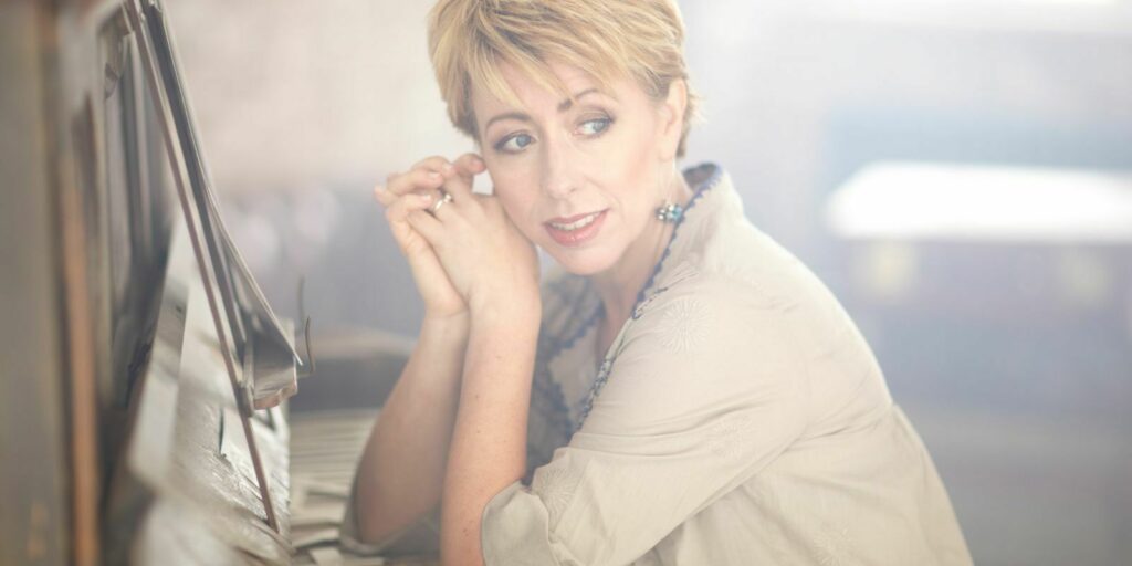Person leaning on a piano and looking at the camera with a gentle expression.