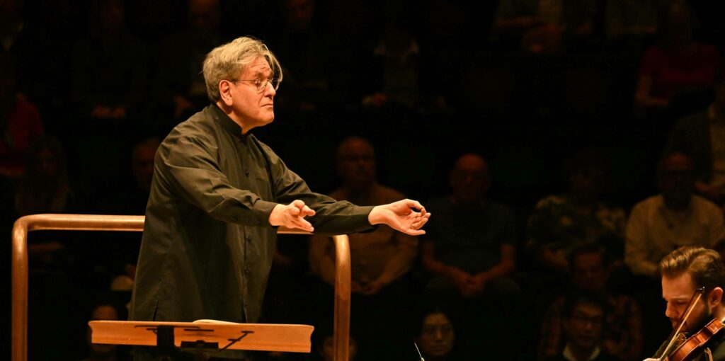 Conductor leads an orchestra performance, standing with arms outstretched in front of music stands, with attentive musicians playing string instruments. Audience visible in the background.
