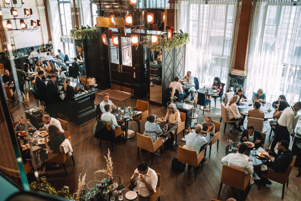 Interior of a busy restaurant with patrons dining at tables, servers attending to customers, and modern décor with large windows and elegant lighting fixtures.