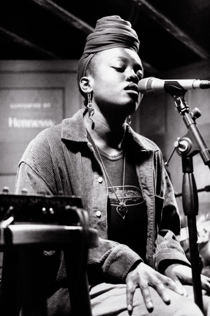 Person singing into a microphone, eyes closed, wearing a large headwrap and a denim jacket. A sign behind reads 'Hennessy.'