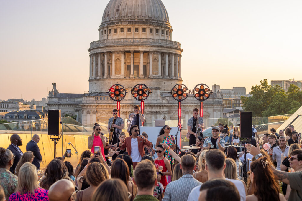 A lively outdoor concert with a band performing in front of St. Paul's Cathedral, surrounded by an enthusiastic audience enjoying the music.
