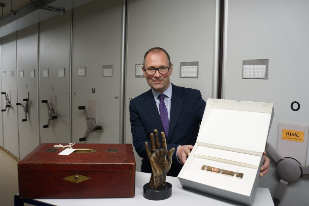 Alen Packwood, Director of the Churchill Archives, sitting at a table displaying objects, including a glove mould and a wooden box, in a room with filing cabinets.