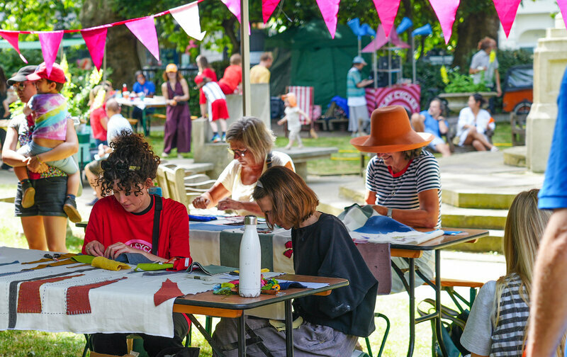 A vibrant fair scene with people of various ages socialising and participating in activities like crafts. Trees and colourful bunting surround the area.