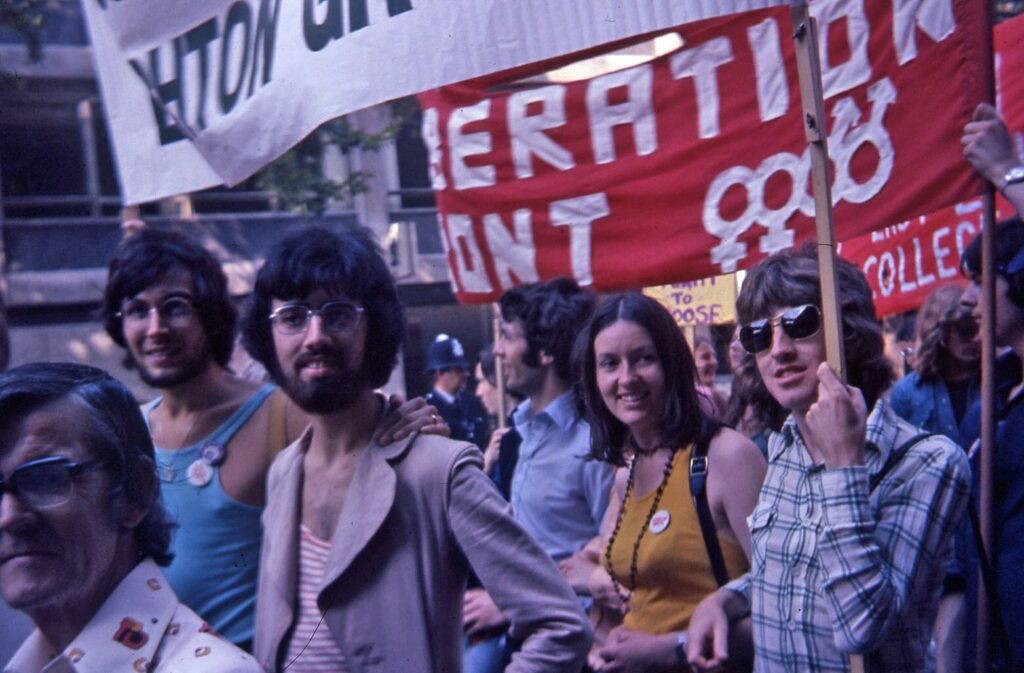 A group of individuals at a protest, holding signs that read "Action for Liberation" and "Vassar College." The atmosphere is lively, with people of various ages and expressions engaging actively with their surroundings.