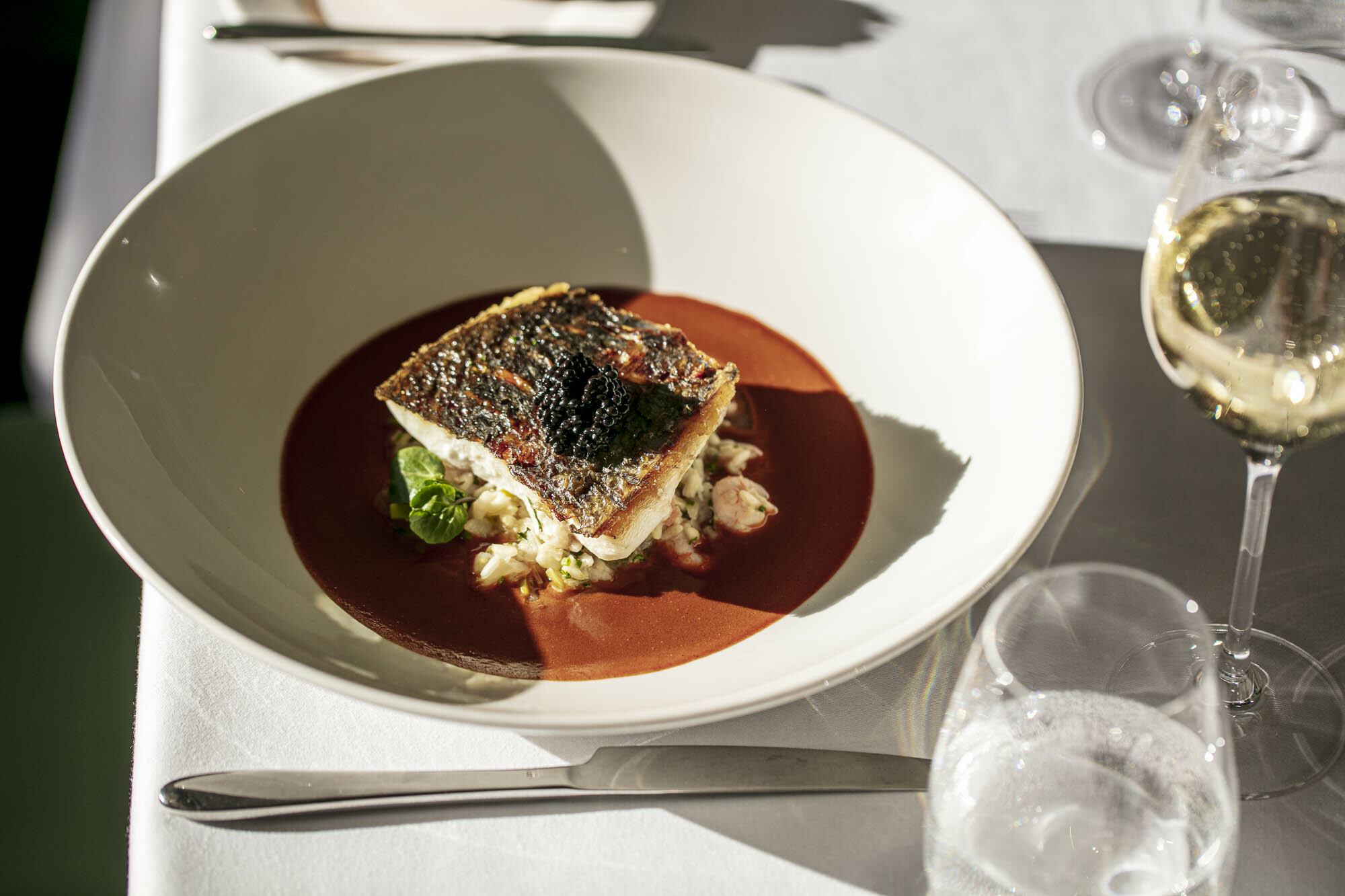 An elegantly plated fish dish topped with caviar is set in a deep red sauce, garnished with greens.