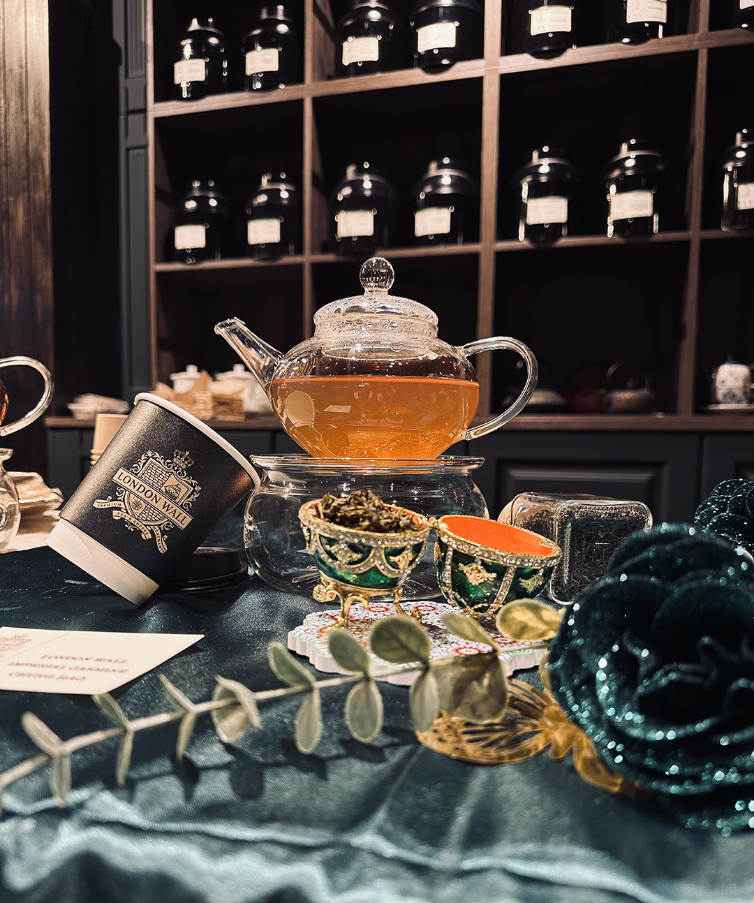 A glass teapot filled with amber liquid is on a table, surrounded by decorative cups and a lush green velvet fabric.